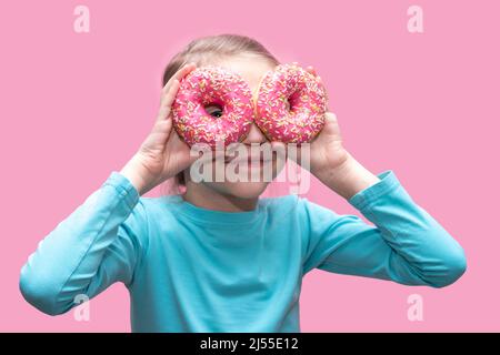 Ein niedliches lustiges Mädchen in einem blauen T-Shirt hält leuchtend rosa Donuts wie eine Brille in der Nähe ihrer Augen und sieht dich auf einem rosa Hintergrund an. Fröhliche Kindheit Konz Stockfoto