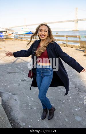 Junge Frau tanzend fröhlich am Pier mit der Oakland Bay Bridge im Hintergrund | Lifestyle Local Tourism Stockfoto