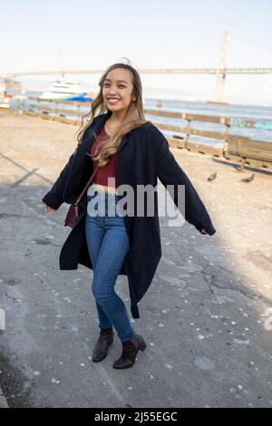 Junge Frau tanzend fröhlich am Pier mit der Oakland Bay Bridge im Hintergrund | Lifestyle Local Tourism Stockfoto