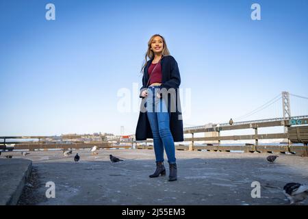 Lächelnde/glückliche junge Frau am Pier mit der Oakland Bay Bridge im Hintergrund | Lifestyle Local Tourism Stockfoto