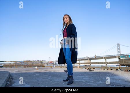 Lächelnde/glückliche junge Frau am Pier mit der Oakland Bay Bridge im Hintergrund | Lifestyle Local Tourism Stockfoto