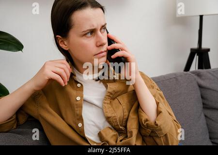 Ängstliche junge Frau, die telefonisch anruft, Smartphone spricht und allein zu Hause schlechte Nachrichten bekommt. Trennung, Stress, Probleme bei der Arbeit oder Beziehungen, Stockfoto