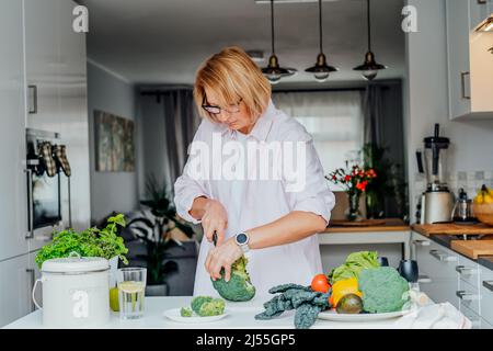 Frau mittleren Alters schneiden Brokkoli beim Kochen gesundes Gericht für das Mittagessen in der Küche. Gesunder Lebensstil. Kochen Zu Hause. Bereiten Sie Essen Zu. Vegetarisch, V Stockfoto