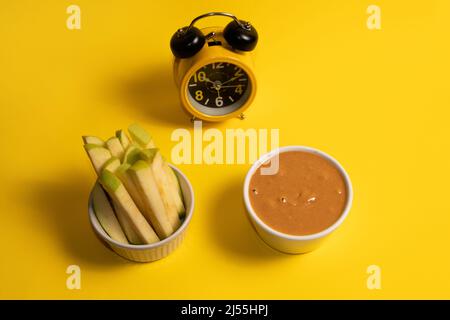 In Scheiben geschnittene ​​apple und Erdnussbutter in Tassen mit Wecker im Hintergrund auf gelbem Hintergrund Stockfoto