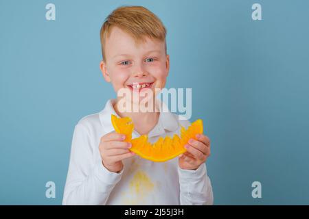 Positiver Junge, der eine geschnittene Hälfte einer Orange hält. Schmutzige Flecken von Orangensaft auf der Kleidung. Das Konzept der Reinigung Flecken auf Kleidung Stockfoto