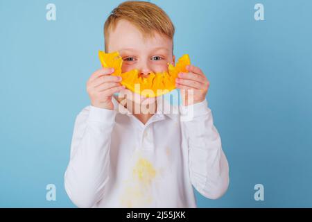 Positiver Junge, der eine geschnittene Hälfte einer Orange hält. Schmutziger Orangensaftfleck auf der Kleidung. Das Konzept der Reinigung Flecken auf Kleidung Stockfoto