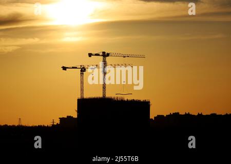 Silhouetten von zwei Turmdrehkranen und unfertigen Gebäuden bei Sonnenuntergang. Wohnungsbau, Wohnblock in der Stadt auf dramatischen Himmel Hintergrund Stockfoto