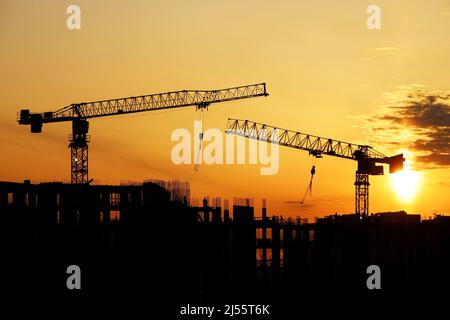 Silhouetten von Baukräne und Arbeitern auf Gerüsten von unfertigen Gebäuden bei Sonnenaufgang. Wohnungsbau, Wohnblock in der Stadt Stockfoto