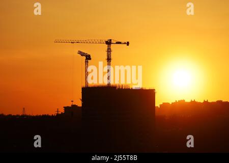 Silhouetten von zwei Turmdrehkranen und unfertigen Gebäuden bei Sonnenuntergang. Wohnungsbau, Wohnblock in der Stadt auf dramatischen Himmel Hintergrund Stockfoto