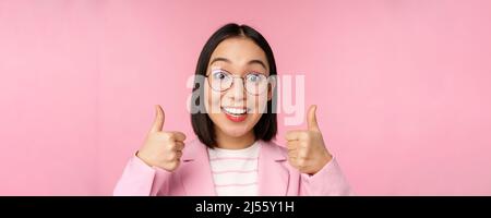 Super, Glückwunsch. Gesicht einer aufgeregten asiatischen Geschäftsfrau in einer Brille, lächelnd zufrieden, mit Daumen nach oben, stehend über rosa Hintergrund Stockfoto