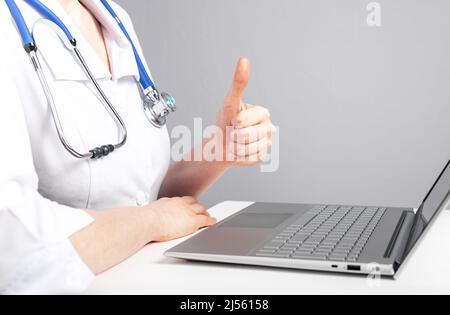 Arzt gibt Online-Konsultation für den Patienten und zeigt Daumen nach oben Geste. Telemedizin, telemedizinisches Konzept. Frau mit Stethoskop sitzt am Tisch mit Laptop und mit Video-Chat. Foto Stockfoto