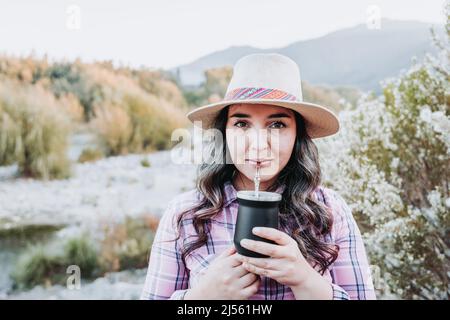 Junge lateinerin mit Hut, trinkt traditionelle Yerba Mate Infusion, in einem natürlichen Raum. Stockfoto