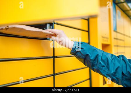 Hand mit Paket. Senden oder empfangen Sie das Paket mit einem gelben Selbstbedienungsautomaten am Postterminal. Stockfoto