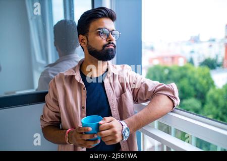 arabischer Mann in legerem Kleid, der eine blaue Tasse auskleidet und traumhaft aussieht Stockfoto