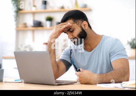 Erschöpfter indischer oder arabischer junger Erwachsener, Freiberufler, Designer, in lässiger Kleidung, sitzen in der Küche, müde von vielen Aufgaben, in Stress, nahm eine Pause von der Arbeit, brauchen eine Pause, haben Kopfschmerzen, Migräne Stockfoto