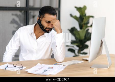Kopfschmerzen und Migräne. Erschöpfter indischer oder arabischer Mann, Makler, finanzieller Mentor, Büroangestellter, der Arbeit müde, In Stress, machte eine Pause von der Arbeit, brauchen eine Pause, bedeckte seine Augen mit den Händen Stockfoto