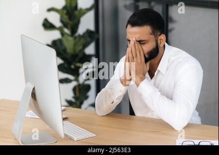 Kopfschmerzen und Migräne. Erschöpfter indischer oder arabischer Mann, Makler, finanzieller Mentor, Büroangestellter, der Arbeit müde, In Stress, machte eine Pause von der Arbeit, brauchen eine Pause, bedeckte seine Augen mit den Händen Stockfoto