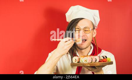 Junger Mann in Gläsern, als Koch gekleidet, hält Holzbrett mit Sushi-Rollen auf rotem Hintergrund. Männliche Köchin isst appetitliche Sushi-Rolle mit Essstäbchen Stockfoto