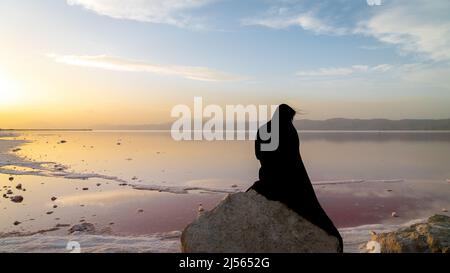 Shiraz, Iran - 2019. Mai: Nicht identifizierte verschleierte iranische Frau im schwarzen Hijab-Kleid, die auf einem Felsen am rosa See von Maharloo sitzt Stockfoto