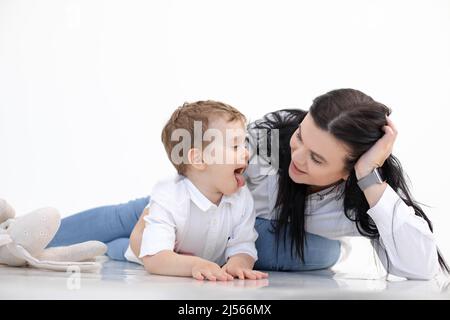 Die schwarzhaarige Lehrerin und der kleine Junge zeigen die Zunge und liegen auf dem Boden. Logopädie, Frühentwicklung, Logopädie Stockfoto