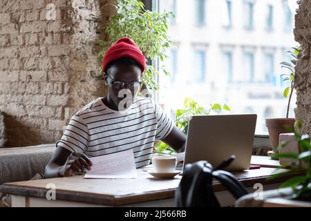 Nachdenklicher afroamerikanischer Hipster-Mann in Brille, der im Café auf einem Laptop mit Dokumenten arbeitet Stockfoto