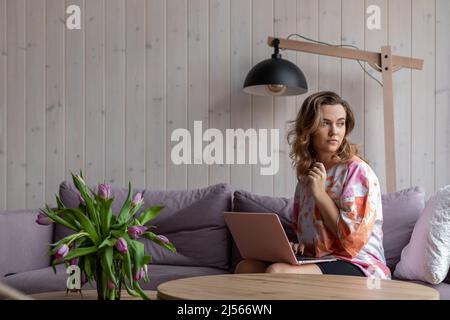 Porträt einer Frau, die auf einem modernen Laptop auf einem weichen, violetten Sofa in legerer Kleidung neben dem Tisch sitzt, mit einer Vase mit ausbleichenden Tulpen. Wohnzimmer in elegant Stockfoto