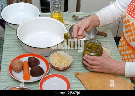 Oben auf dem Kochtisch mit der Hand einer Frau, die eingelegte Gurken aus dem Glas in der Nähe von Geschirr und Gemüse für das Kochen von Salat arrangiert. Stockfoto