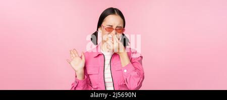 Stinkend. Asiatisches Mädchen in Sonnenbrille und stilvollem Outfit, schließt ihre Nase vor Ekel, mag schlechten Geruch nicht und steht über rosa Hintergrund Stockfoto