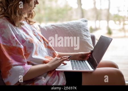 Nahaufnahme einer jungen Frau, die auf einem modernen Laptop tippt und arbeitet und auf einem weichen Sofa in legerer Kleidung in der Nähe eines großen Panoramafensters sitzt. Wohnzimmer Stockfoto