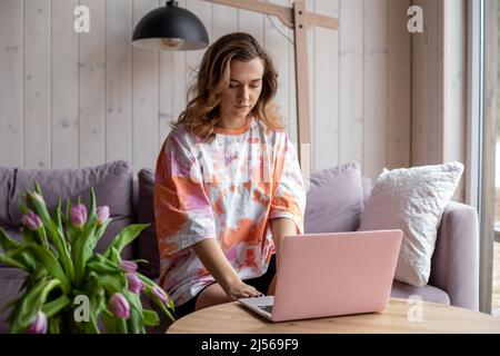 Porträt einer Frau, die auf einem modernen rosa Laptop tippt und in legerer Kleidung auf einem weichen Sofa neben dem Tisch sitzt, mit einer Vase aus verwelkenden Tulpen. Wohnzimmer in Stockfoto