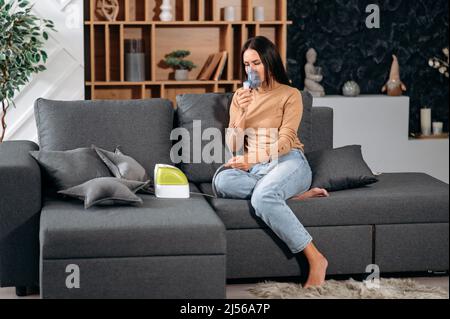 Krank verärgert kaukasische Brünette Frau mit einem Inhalator, krank ungesunde Frau beim Inhalieren zu Hause, sie verwenden Vernebler und Inhalator für die Behandlung, sitzen auf der Couch zu Hause, brauchen Behandlung Stockfoto