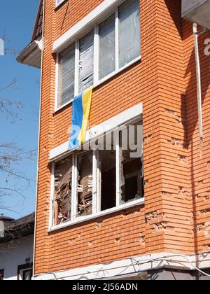 Zerstörte Gebäude nach dem Bombenanschlag, Nahaufnahme. Ukrainische Flagge hängt auf dem Balkon eines geschälten Hauses in der Ukraine. Beschuss von Wohngebäuden mit Minen. Befreite ukrainische Stadt von russischen Eindringlingen Stockfoto