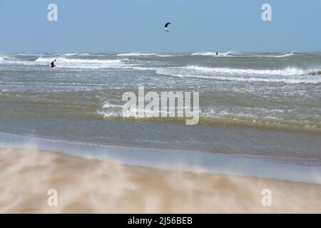 Starke Winde wehen Sand über den Strand, während ein Kite-Surfer reitet und ein Paar in der schweren Brandung spielt. South Padre Island, Texas. Stockfoto