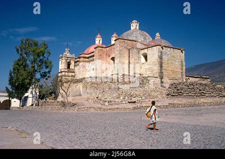 Ein Mädchen aus Zapotec überquert eine gepflasterte Straße mit Blick auf die alte spanische Mission in Mitla, einer 1000 Jahre alten Stadt in Zapotec in der Nähe von Oaxaca in der südlichen Sierra Madre, Mexiko.die Kirche ist die Templo de San Pablo Apostol befindet sich innerhalb der archäologischen Stätte von Mitla, Etwa 46 km (29 mi) von Oaxaca-Stadt entfernt. Stockfoto