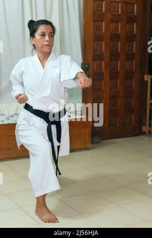 Hispanische Frau in traditionellem Kimono, die zu Hause Karate praktiziert - Schwarzer Gürtel Stockfoto