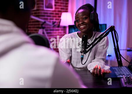 Weibliche Vlogger interviewt männlichen Gast auf Podcast-Show, mit Aufnahme-Ausrüstung. Glückliche Frau streamt Live-Broadcast-Episode mit Mann im Studio, um Gespräch für Kanal aufzunehmen. Stockfoto