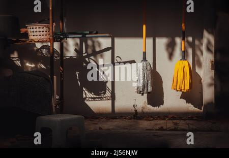 Pinkfarbener Wäschekorb aus Kunststoff und zwei Wischmopps, die nach der Reinigung durch Sonnenlicht an der Betonwand hängen. Hausreinigungskonzept BAC Stockfoto