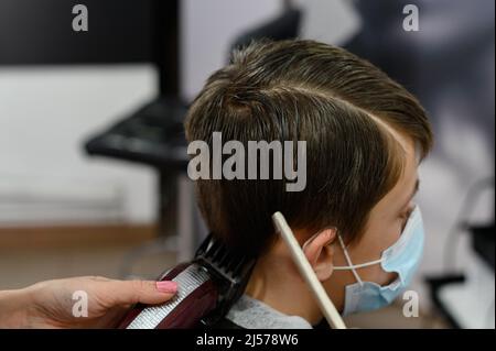 Ein Teenager in einem Schönheitssalon bekommt einen Haarschnitt, ein Friseur schneidet den Jungen eines Teenagers, einen Haarschnitt mit einem Haarschneider und einem Kamm. Stockfoto