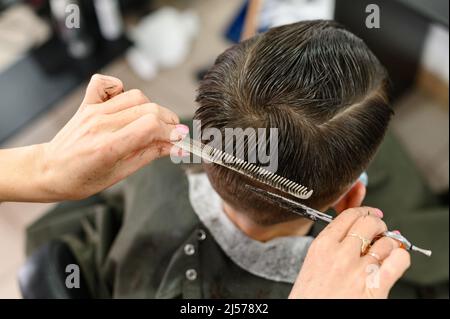 Teen Kerl bekommt einen Haarschnitt während einer Pandemie in einem Friseurladen, Haarschnitt und Trocknen der Haare nach einem Haarschnitt, Haarschnitt mit einer Schere. Stockfoto