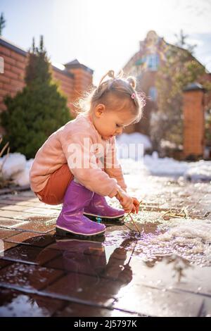 Mädchen spielt in einer Pfütze mit Holzstock am Frühlingstag bei Sonnenlicht Stockfoto