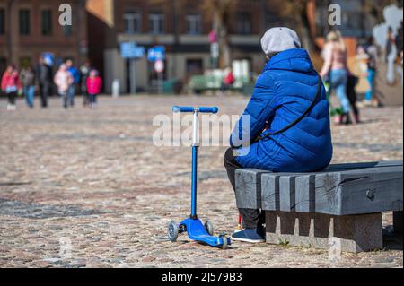 Eine alte Frau ruht auf einer Bank und wartet darauf, dass ihr Enkelkind auf dem Stadtplatz spielt, Rückansicht Stockfoto