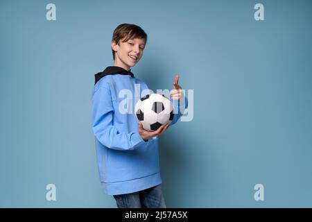 Lächelnder Teenager-Junge in sportblauer Kleidung in einem Sweatshirt, der den Fußballball im Studio auf einem farbigen Hintergrund posiert. Kaukasischer Teenager Fan des Fußballspiels Stockfoto