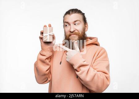 Scandinavian handsome Mann hält Geschenk und zeigt isoliert auf grauem Hintergrund zu präsentieren. Kerl mit Ingwer-Frisur und Bart in einem Sweatshirt. Konzept Stockfoto