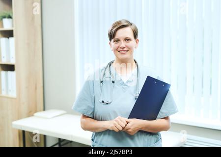 Porträt einer lächelnden jungen kaukasischen Krankenschwester mit Stethoskop, das im Krankenhausbüro mit Zwischenablage steht Stockfoto