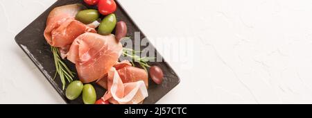 Banner, Draufsicht auf schwarzem Charcuterie-Brett mit Proscuitto serviert mit Oliven und Babytomaten auf weißem Beton-Tischhintergrund mit Kopierfläche. B Stockfoto