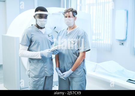 Portrait von multiethnischen MRT-Spezialisten in Scrubs und Masken, die mit einem Tablet gegen ein Ultraschallgerät stehen Stockfoto