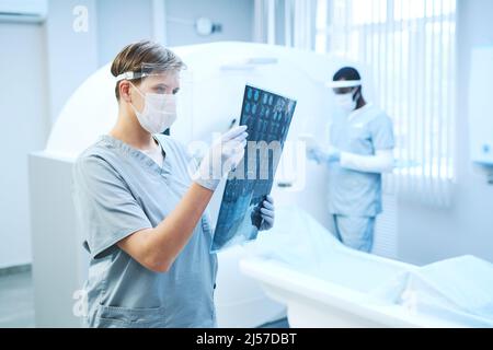 Fokussierter Radiologe in der Sicherheitsabschirmung, der im Krankenhaus mit einem Röntgenbild des Gehirns gegen einen CT-Scanner arbeitet Stockfoto