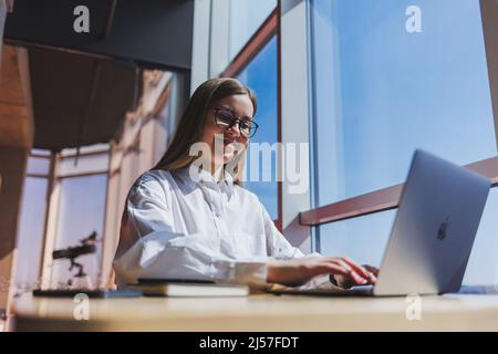 Blick auf eine fokussierte Frau in Freizeitkleidung, die auf Netbook tippt, während sie an einem neuen Projekt am Tisch im modernen Arbeitsbereich sitzt Stockfoto