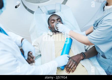 Direkt über der Ansicht von Ärzten, die dem schwarzen Patienten eine Sauerstoffmaske mit geschlossenen Augen auf dem Untersuchungstisch geben Stockfoto