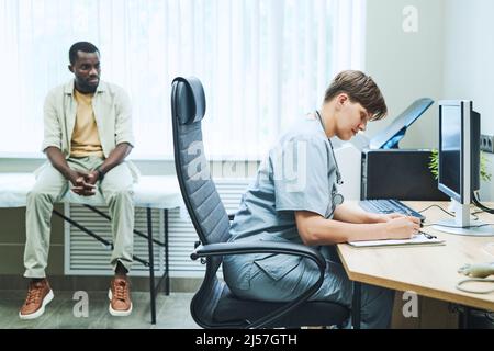 Konzentrierter junger kaukasischer Chirurg, der am Tisch mit Computer sitzt und die Patientenkarte bei der Verabredung füllt Stockfoto